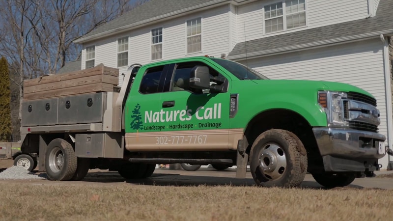 Nature's Call Landscaping branded green Ford pickup truck with stake-body trailer parked on a North Wilmington, Delaware jobsite, Landscape, Hardscape, Drainage signage on the door, phone number 302-777-7767.