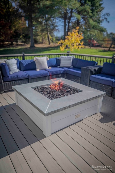 Vertical cinematic composition of the Chadds Ford fire pit: a square white concrete-topped propane fire pit with lit lava rocks centered on the gray composite deck, the curved deep blue wicker sectional behind, and a single yellow-foliage autumn tree silhouetted against the sky