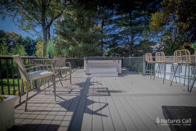 Wide deck-level view of the Chadds Ford hot tub zone: the recessed hot tub at center frame ringed by a small chair seating area on the left and the outdoor kitchen bar with wicker stools on the right, all on the gray composite deck under autumn trees