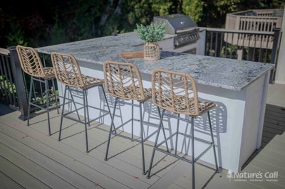 Wide deck-level view of the Chadds Ford outdoor kitchen bar: a granite-topped concrete bar with a small potted herb at center holds four woven wicker bar-height stools in profile, with the stainless gas grill and a section of black aluminum railing visible behind on the gray composite deck