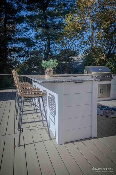 Sun-streaked portrait of the Chadds Ford outdoor kitchen: four wicker bar-height stools line the granite-topped bar, with the gas grill and storage cabinetry visible at the right and a small potted plant on the counter