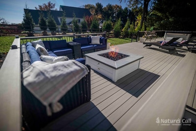 Wide cinematic ground-level view of the Chadds Ford back deck on a clear autumn day: a square white propane fire pit at center frame, ringed by a curved deep blue wicker sectional with cream pillows on the left, two wicker chaise lounges on the right, and the home's tan siding visible at the far edge