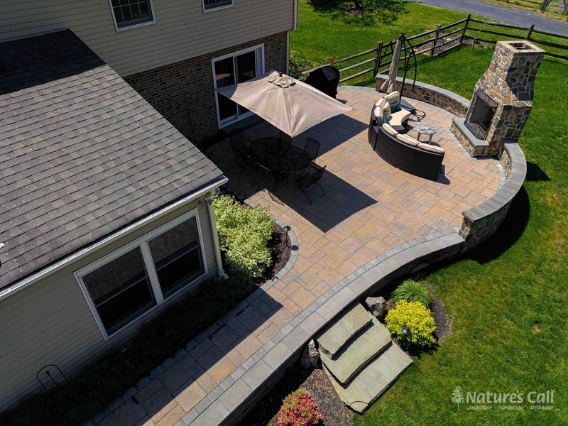 Aerial view of a Garnet Valley Pennsylvania back patio: a curved paver platform surrounds an in-ground hot tub and full-height fieldstone outdoor fireplace, with a tan umbrella shading the dining area, Nature's Call landscaping and hardscaping project