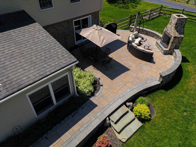 Aerial view of a Garnet Valley PA back patio: a curved paver platform surrounds an in-ground hot tub and full-height fieldstone outdoor fireplace, with a tan umbrella shading the dining area and a low stone retaining wall tracing the patio edge against the lawn beyond