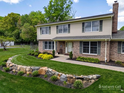 Two-story brick and cream-siding home in Garnet Valley PA viewed from the front lawn. A curved paver walkway with charcoal border bricks leads from the driveway to the columned front entry, flanked by mounded yellow shrubs. A low fieldstone retaining wall edges the lawn in the foreground