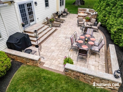 Multi-level paver patio with dining and lounge zones, stone retaining walls, stair access, hedge screening, and outdoor grill.