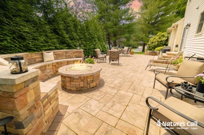 Expansive paver patio with circular stone fire pit, curved seat wall, and multiple lounge seating areas nestled among mature landscaping.