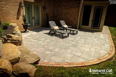 Curved paver patio with timber border, lounge chairs, boulders at foreground, foundation plantings, and brick home entry doors.