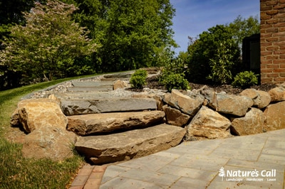 Stone seat wall with bluestone coping, oversized boulder accents, flagstone walkway leading through property with mature shade trees and foundation plantings.
