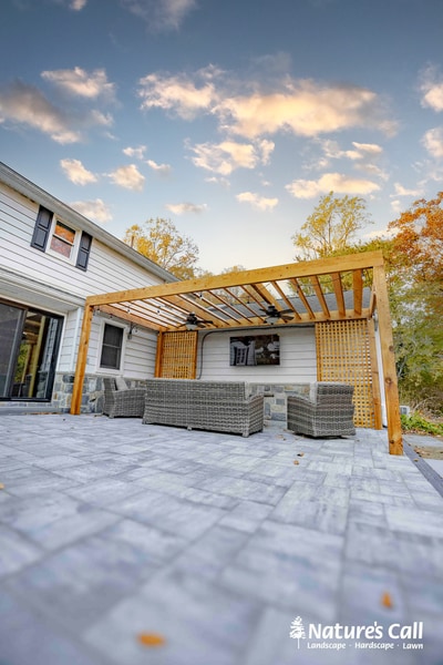 Light gray paver patio with woven rattan outdoor seating beneath a wooden pergola attached to a white modern house with lattice panels.