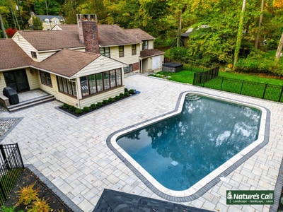 Expansive paver patio with white border accents surrounding a blue swimming pool, adjacent to a modern home with enclosed sunroom and metal railing.