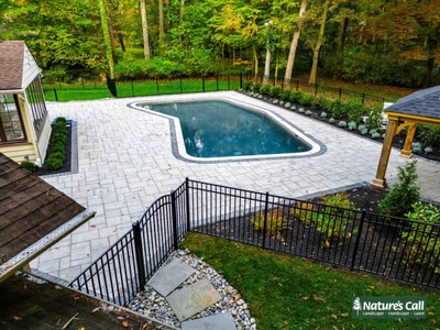 Light gray paver patio with integrated pool surround, blue swimming pool with white border, black metal railing, and woodland backdrop with young plantings.