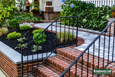 Curved brick steps with black metal railings and planting beds border a bluestone walkway between home and landscaped gardens.