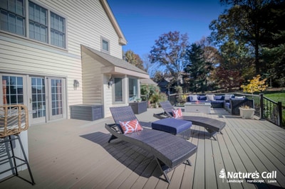 Large composite deck with black metal railings and lounge seating extends from modern home exterior with mature landscape backdrop.