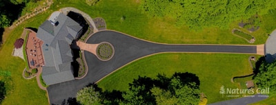 Top-down drone view of the entire Hockessin estate property: the long driveway sweeps in a curve from the side, narrows past the multi-gable brick home at the upper left, and opens into a circular paver loop with the boxwood-planted oval centerpiece, framed by mature woodland and a small entry walkway tucked alongside the home