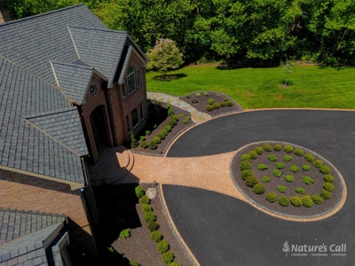 Aerial three-quarter view of the Hockessin estate showing the brick paver entry path tucked between the home and the boxwood-planted oval centerpiece, with mounded boxwoods framing the side and a curved cobble border defining the bed edge