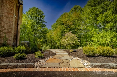 Wide ground-level view of the Hockessin side path: irregular flagstone steppers cross a river-rock drainage strip leading away from a brick step landing, with mounded boxwoods, a dogwood in bloom, and a woodland edge framing the approach beyond