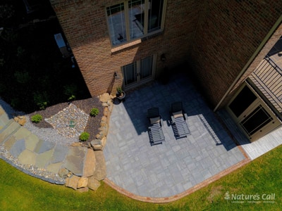 Aerial of the Hockessin back patio: a small gray paver patio with two chaise lounges sits tucked against the brick home's exterior corner, edged by a brick band and a flagstone walkway with rough boulder borders