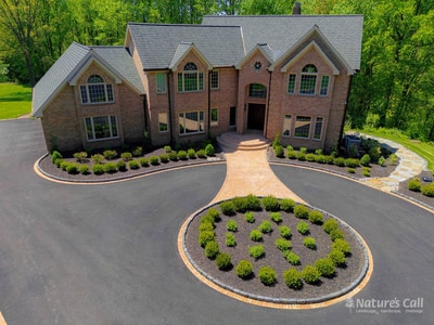 Hockessin Delaware estate after Nature's Call rebuild: brick paver walkway and entry, fresh charcoal asphalt driveway with brick borders, brick-edged boxwood-planted oval centerpiece, and refreshed mulched foundation beds with manicured boxwoods.