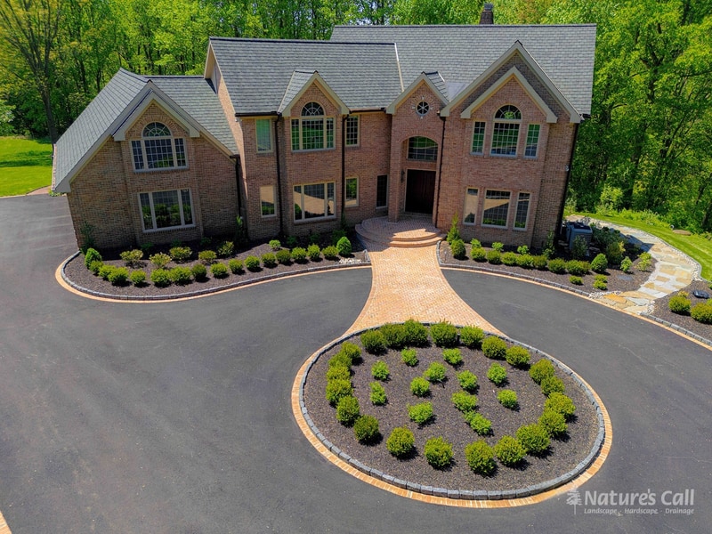 After, Hockessin estate brick home with rebuilt brick paver walkway and entry, fresh charcoal asphalt driveway with brick borders, brick-edged boxwood-planted oval centerpiece, and refreshed mulched foundation beds with manicured boxwoods, Nature's Call Landscaping