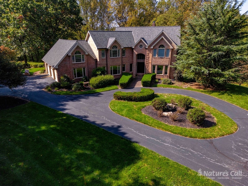 Before, Hockessin estate brick home with original asphalt circular driveway, an oval center bed of loose stones and patchy plantings, and basic foundation shrubs along the front of the house, Nature's Call Landscaping
