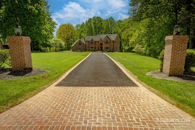 Long view up the Hockessin driveway from the road: a brick paver runner stretches between two brick gate columns with lantern caps, a smooth lawn extends on both sides, and the brick estate sits centered at the end of the approach