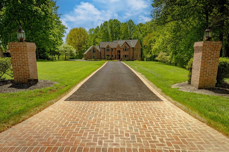 Hockessin Delaware estate approach: a brick-bordered driveway runs up a broad, manicured lawn graded to shed water, flanked by brick lamp pillars, with the brick home set among mature trees, Nature's Call landscape, hardscape, and drainage project
