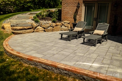 Detail view of a Hockessin planting bed edge: a low stacked-stone wall in dark gray fieldstone separates a mulched bed of mounded boxwoods from the asphalt drive, with a brick paver runner curving past at the right edge