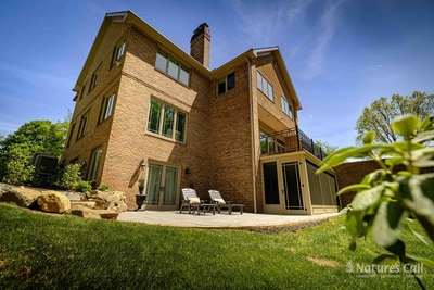Wide ground-level view of the back of the Hockessin estate: the tall brick facade with chimney rises across the frame, a small back patio with a pair of chaise lounges sits below the walkout level, and a smooth green lawn extends to the woodland edge at the right