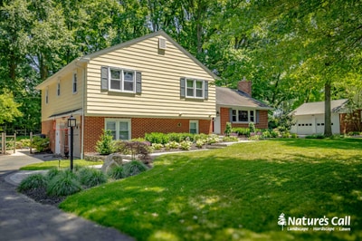 Mid-century brick and siding home features curved driveway with ornamental grasses, perennial beds, and mature shade trees defining the entry.
