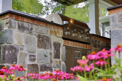 Close-up of the North Wilmington outdoor kitchen: the stone-faced bar with a stainless gas grill in focus, copper-toned tile backsplash, a small smoker on top of the grill, and a row of pink coneflower blooms soft-focus in the foreground