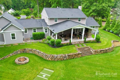 Aerial three-quarter view of the North Wilmington back yard: the covered timber pavilion at center, the curved dry-stack fieldstone retaining wall sweeping from left to right, hand-cut stone tread steps connecting the lawn to the patio level, and the round dry-stack fire pit visible in the lawn at lower left