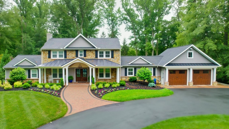 Front view of a North Wilmington Delaware home with stone-and-cedar-shake facade, a curved tan paver driveway, and layered foundation plantings of liriope, boxwoods, hydrangeas, and seasonal annuals, Nature's Call landscape, hardscape, and outdoor living project