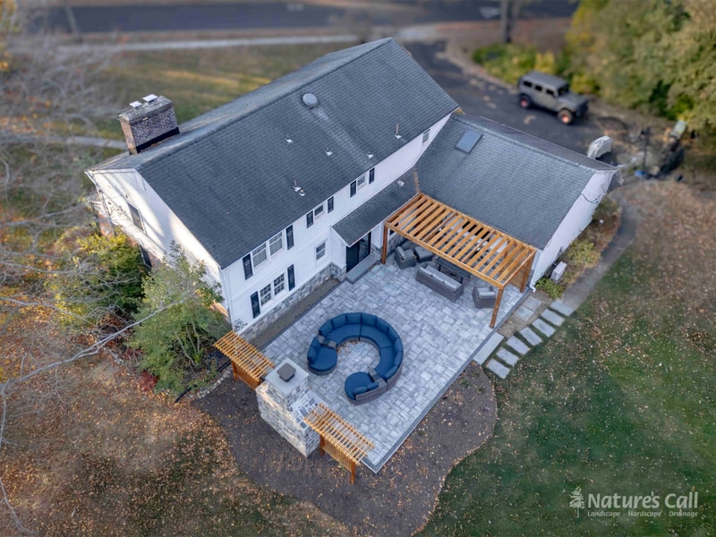 Aerial dusk view of a North Wilmington Delaware property after Nature's Call rebuilt the back yard: a new gray paver patio with curved wicker sectional surrounds a stand-alone fieldstone outdoor fireplace, and a cedar pergola shades the patio against the home, Nature's Call hardscape and outdoor living project.