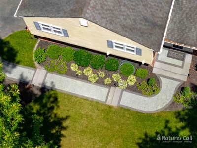 Aerial view of a paver front walkway and entry with a darker stone border, flanked by layered foundation plantings of hostas, shrubs, and perennials along a tan-sided home. Nature's Call Landscaping hardscape project.