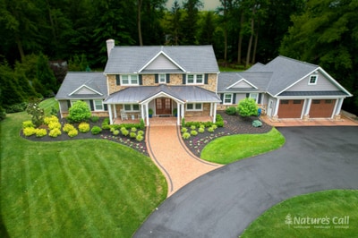 Aerial view of a North Wilmington property with a stone-and-cedar-shake home: a curved brick paver driveway with charcoal Belgium-block edge sweeps to the front entry, and layered foundation plantings of mounded shrubs and seasonal annuals trace the front of the house. Nature's Call Landscaping in North Wilmington.