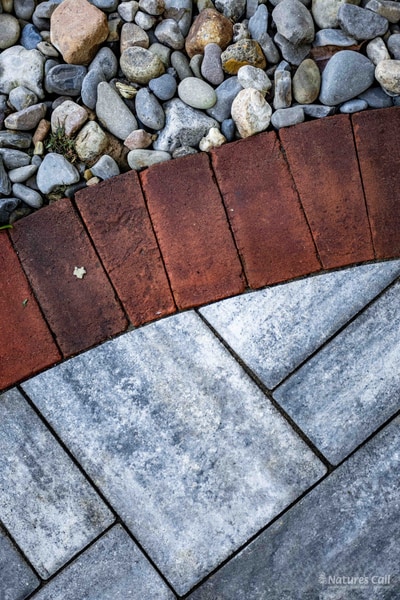 Vertical material macro at the Wilmington alley edge: blue-gray pavers in the foreground meeting the red brick curb, with a strip of river-rock drainage stones beyond, three of the project's core hardscape materials in one frame