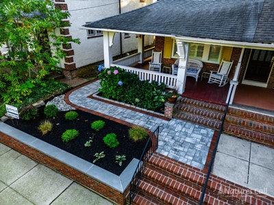 Aerial three-quarter view of the Wilmington front entry: the covered porch with white wicker chairs sits at center, the curved paver walkway with red brick edge sweeps in from the sidewalk, and the bluestone-capped brick planter beds hold hydrangeas and mounded boxwoods