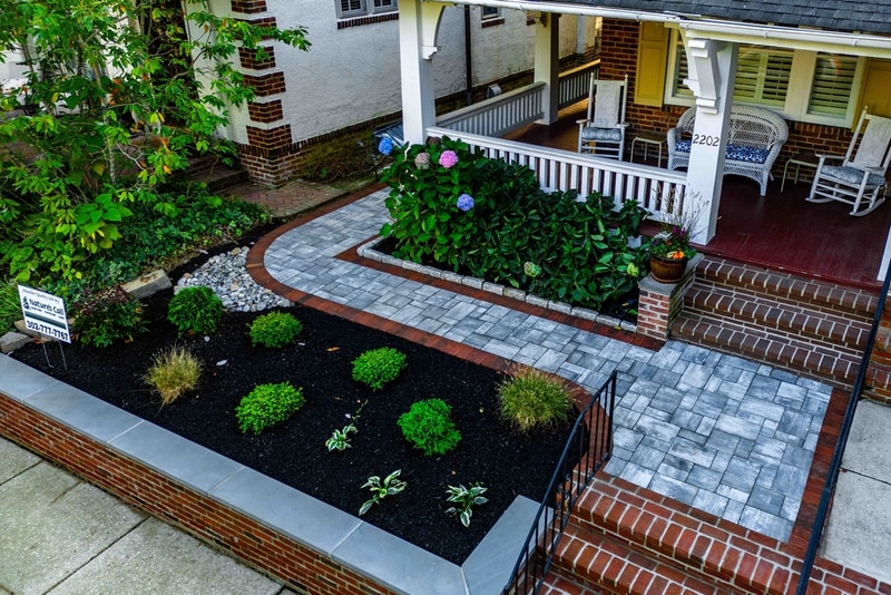 Aerial three-quarter view of the Wilmington front-yard hardscape: a curved blue-gray paver walkway with red brick edging sweeps from the sidewalk to the porch, a hedge of pink and blue hydrangeas anchors the planter bed center, brick-faced raised planters with bluestone caps frame both sides, mounded boxwoods and ornamental grasses fill the mulch bed at left, and brick risers rise to the paver landing at the porch on the right
