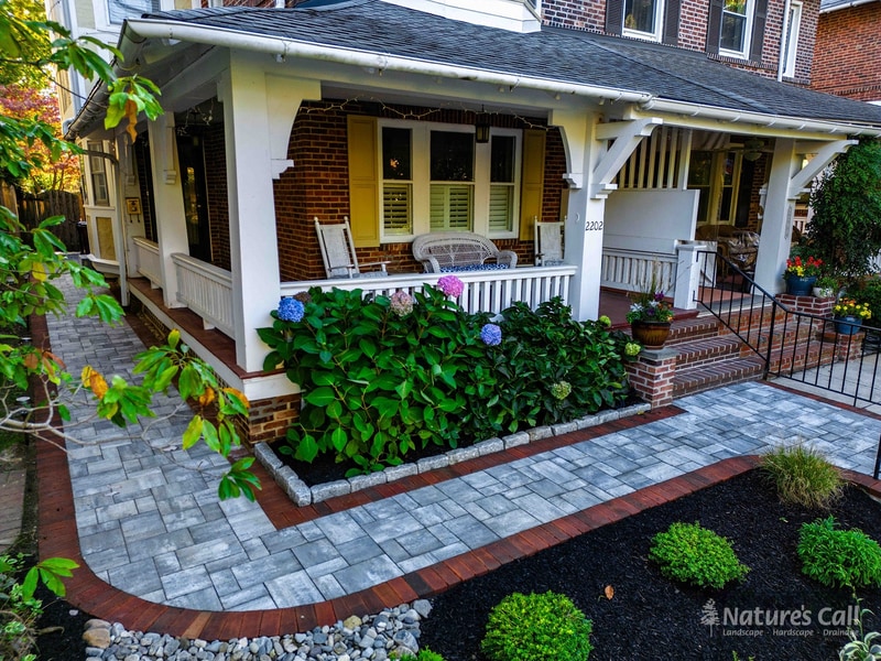 Front-yard view of a Wilmington Delaware row home with curved blue-gray paver walkway, red brick edging, brick-faced raised planters, and a hedge of blue and purple hydrangeas, Nature's Call hardscaping and front-entry rebuild